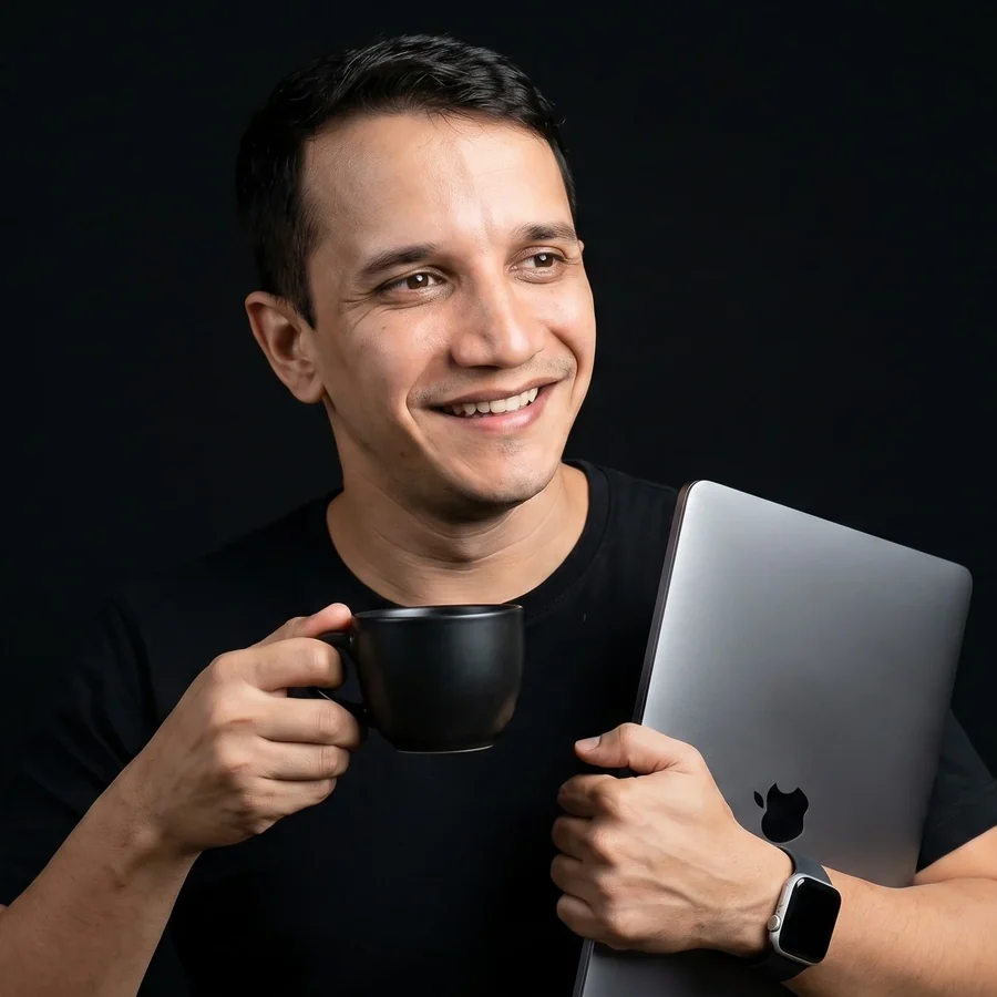 Djean Moraes em pé, segurando uma xícara em uma das mãos e um notebook fechado debaixo do braço, em fundo escuro